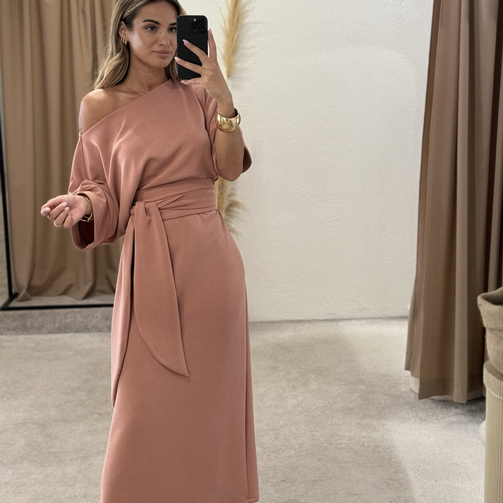 model wearing off shoulder blush dress with belt in dressing room model wearing off shoulder blush dress with belt in dressing room
