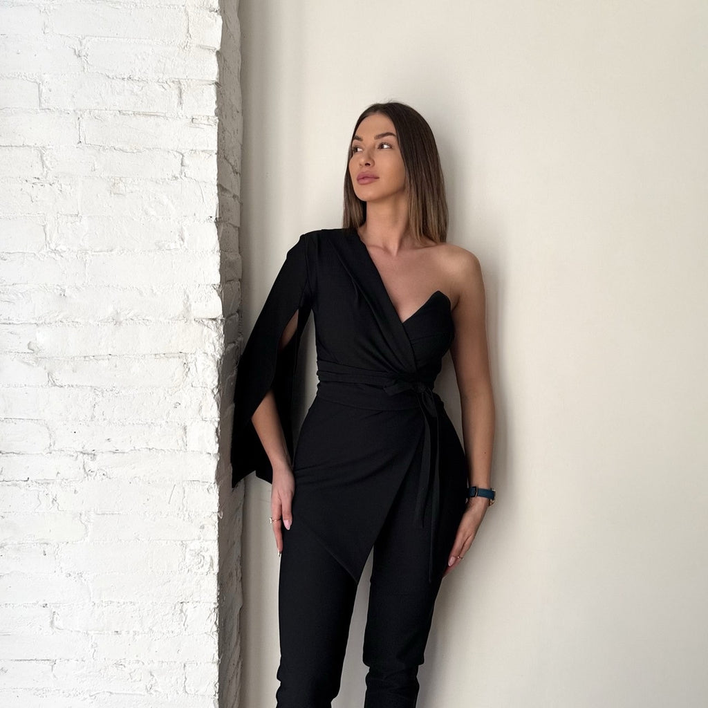 woman in black one shoulder jumpsuit standing against white brick wall woman in black one shoulder jumpsuit standing against white brick wall