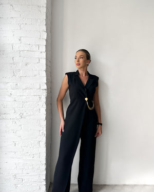 woman wearing black jumpsuit with chain belt standing against white brick wall