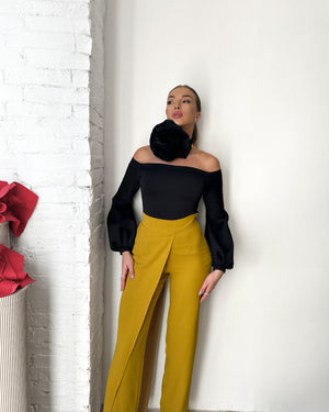 woman in black top and yellow pants standing against white wall